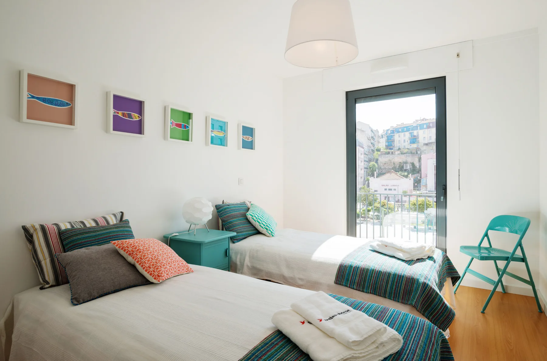 A bright bedroom with two beds, colorful cushions, and a glass door opening to a balcony.