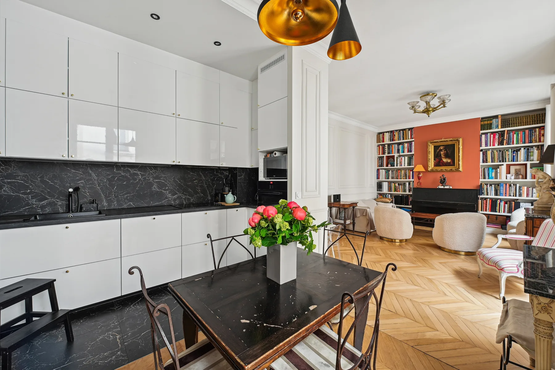 Un comedor luminoso con una mesa negra, armarios blancos y una zona de estar con estanterías.