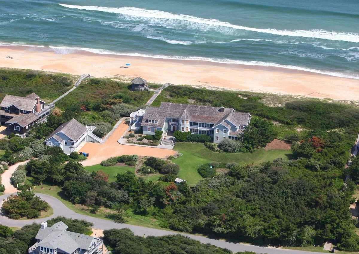 Vista aérea de casas junto al mar entre vegetación, con una playa de arena y el océano al fondo.