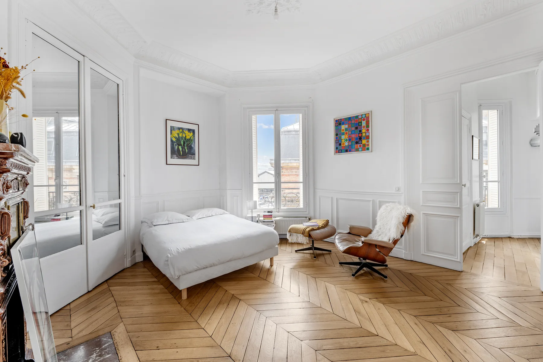A bright bedroom with a bed, a pair of lounge seats, and framed artwork.