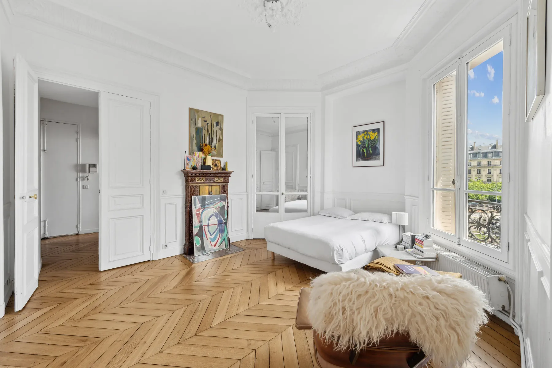 A bright bedroom with a white bed, fireplace, and tall windows overlooking the city.
