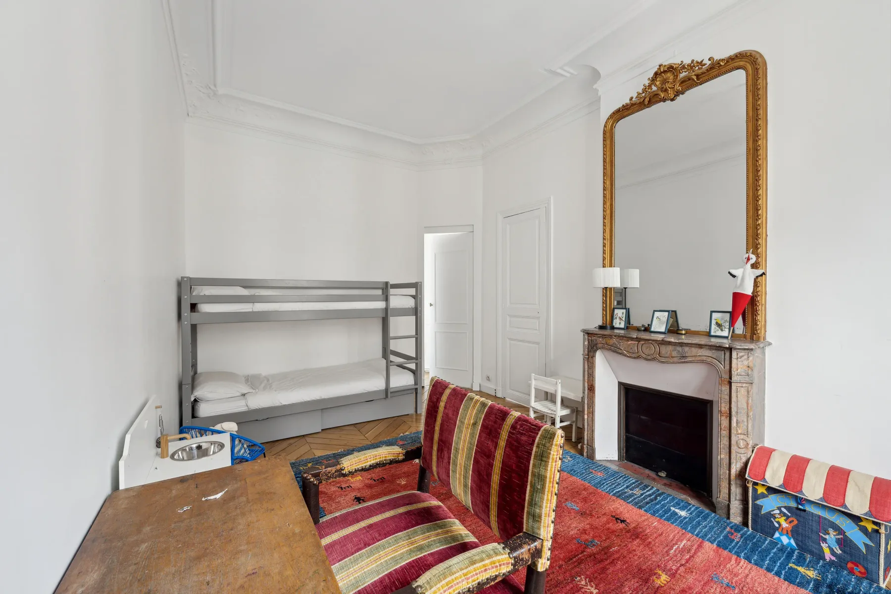 A bright bedroom with bunk beds, colorful seating, and an ornate fireplace.