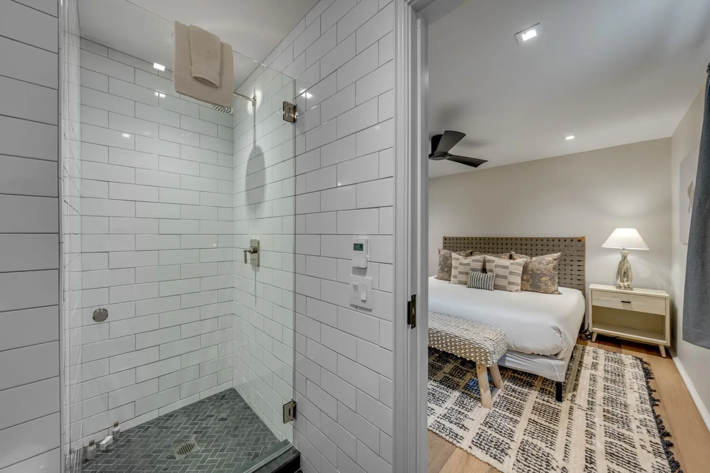 A tiled shower opens onto a bedroom with a bed and patterned rug.