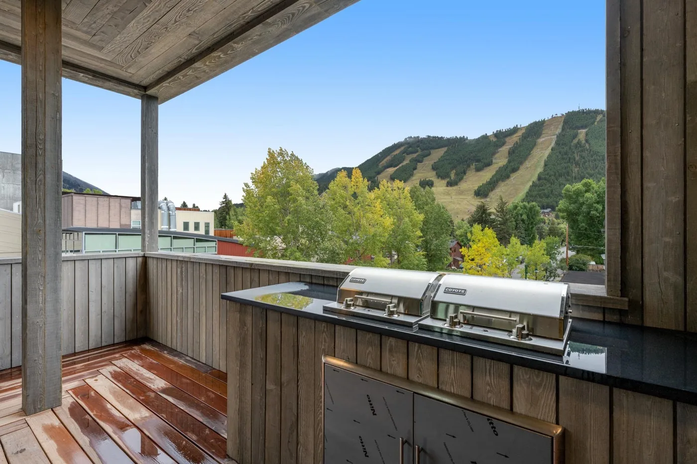 A covered outdoor deck with a built-in grill overlooks trees and a green hillside.