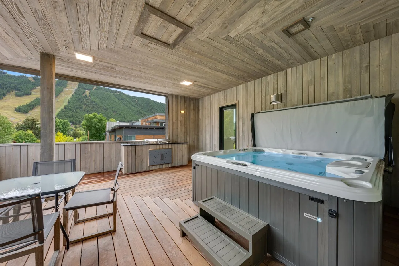 A covered deck features a hot tub, outdoor seating, and a view of green hills.