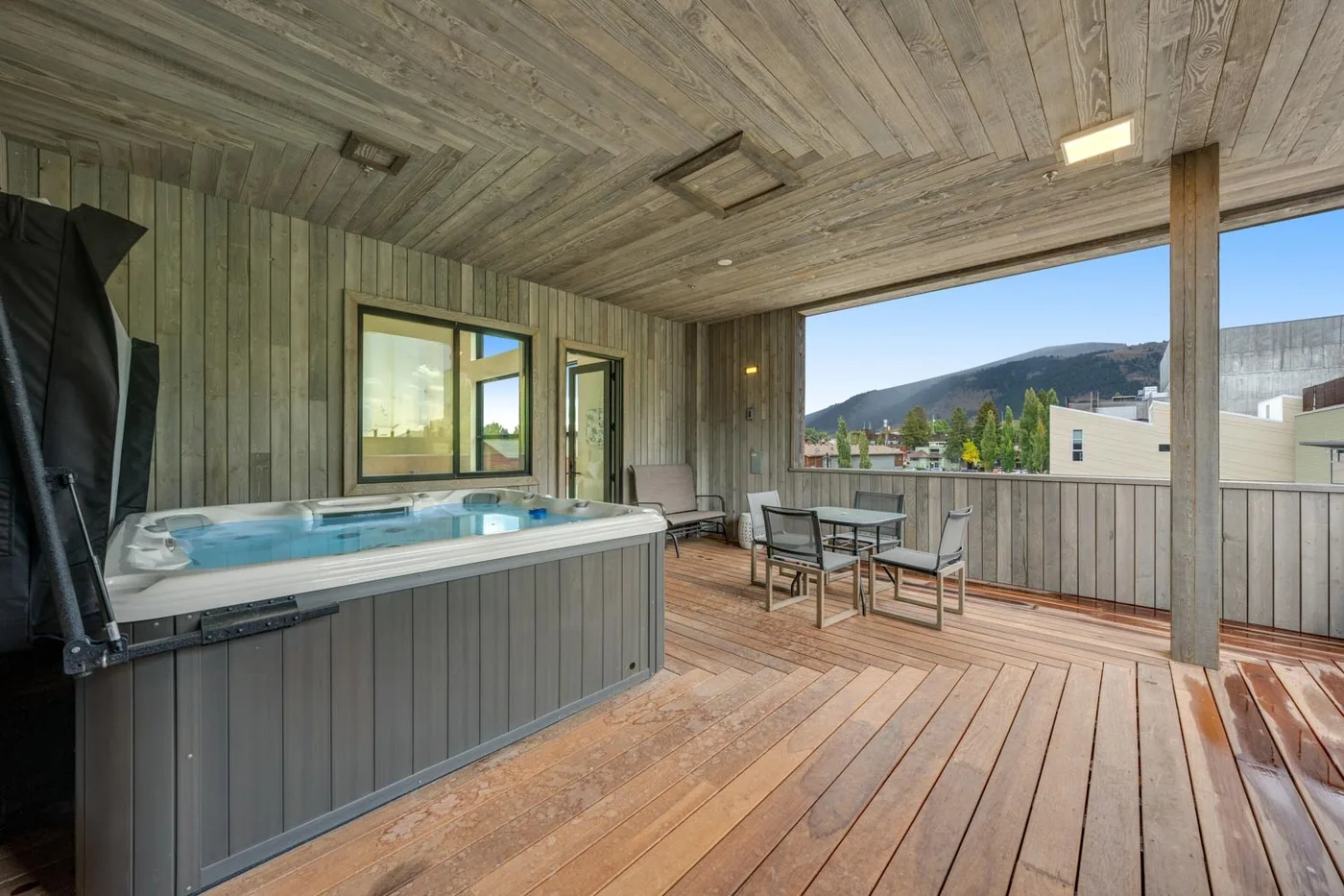 A covered wooden deck with a hot tub and seating overlooks a mountain view.
