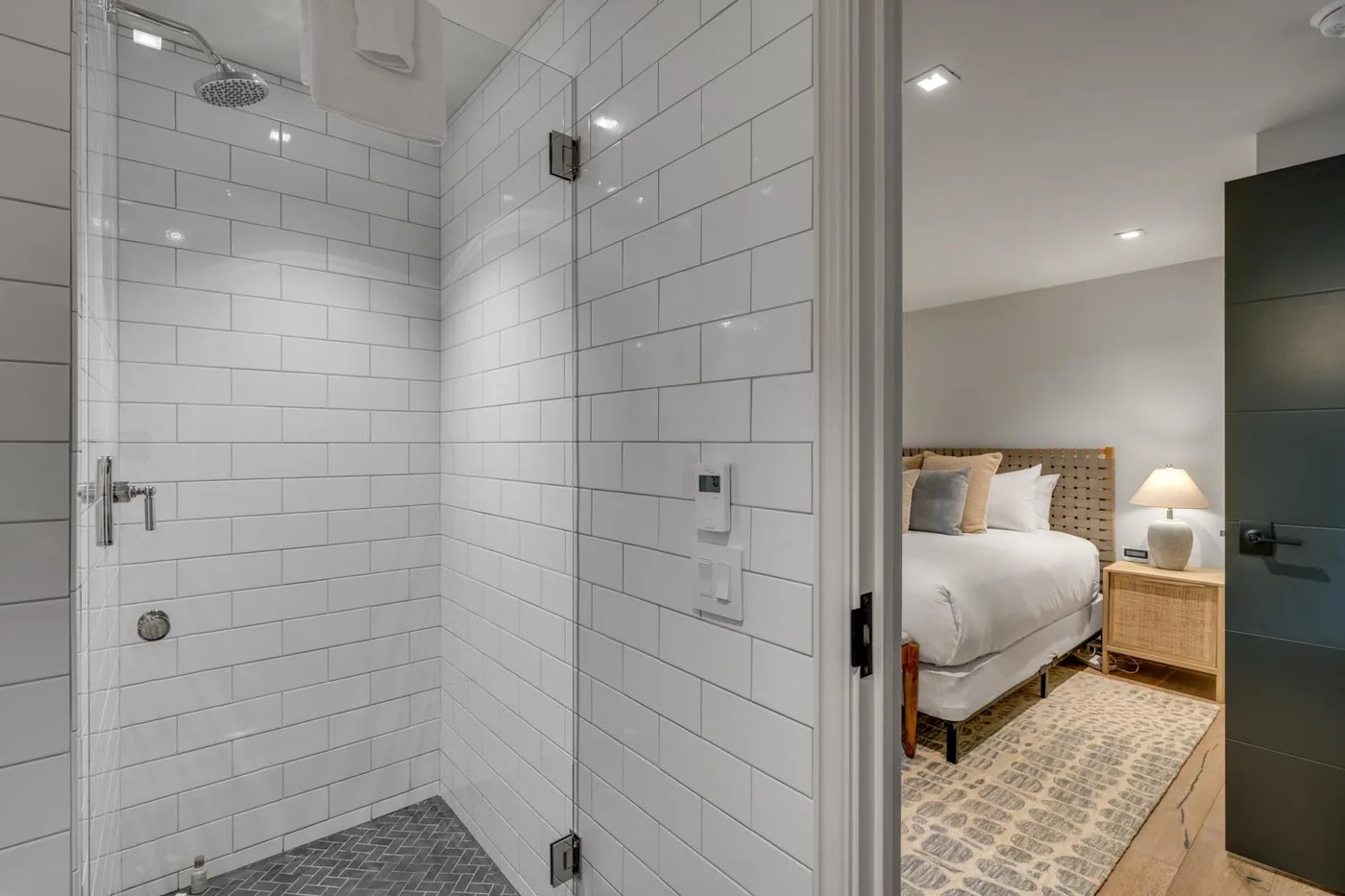 A tiled bathroom shower opens onto a bedroom with a bed and bedside table.