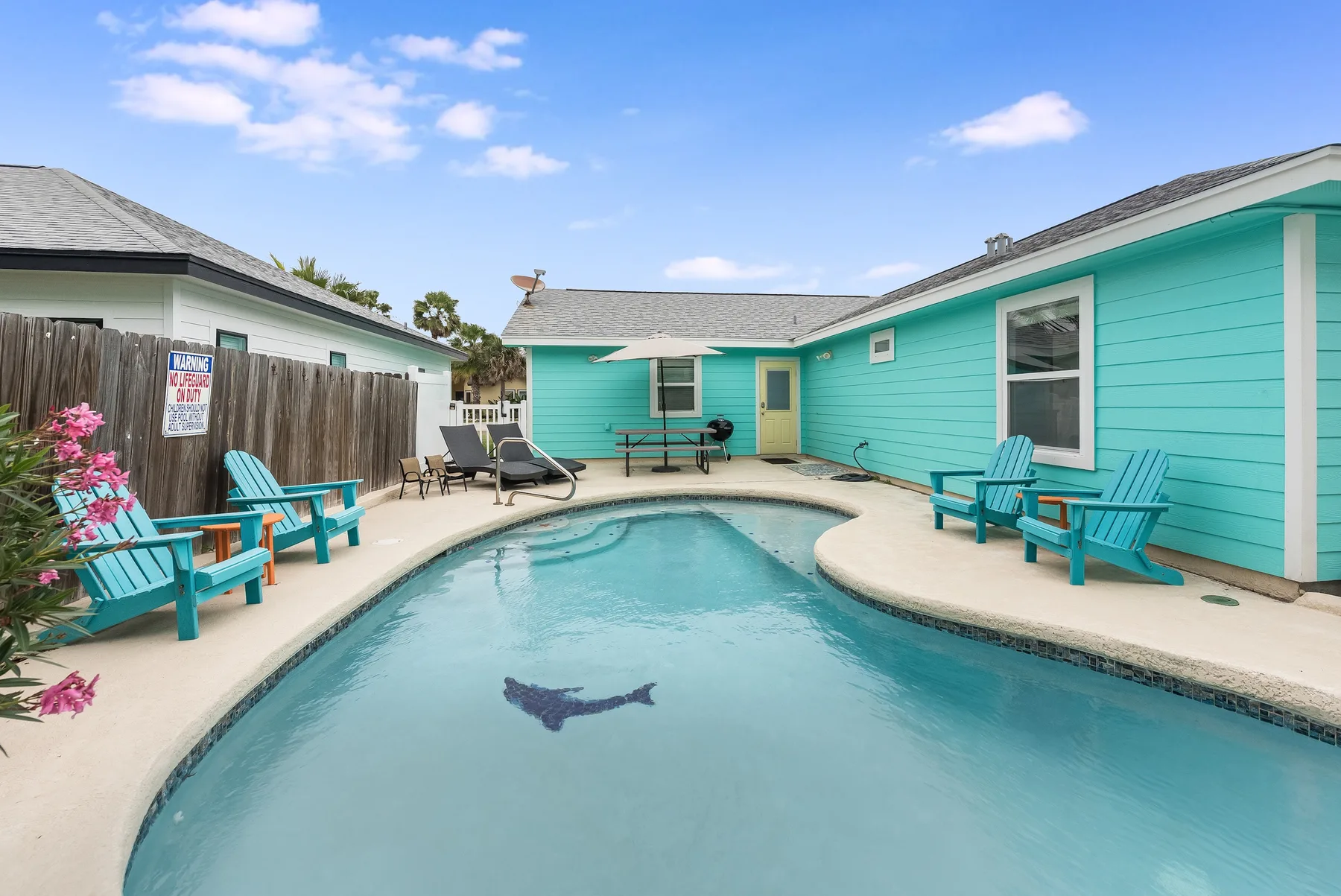 A swimming pool sits in a courtyard beside turquoise buildings and outdoor seating.