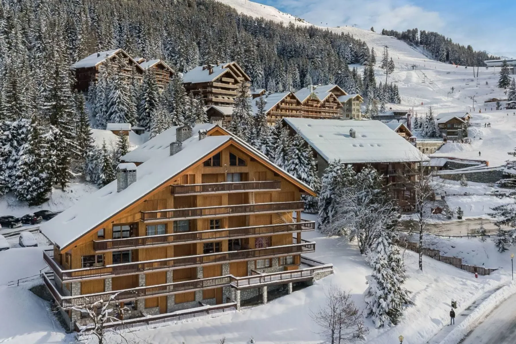 Edificios tipo chalet cubiertos de nieve junto a una pista de esquí boscosa.
