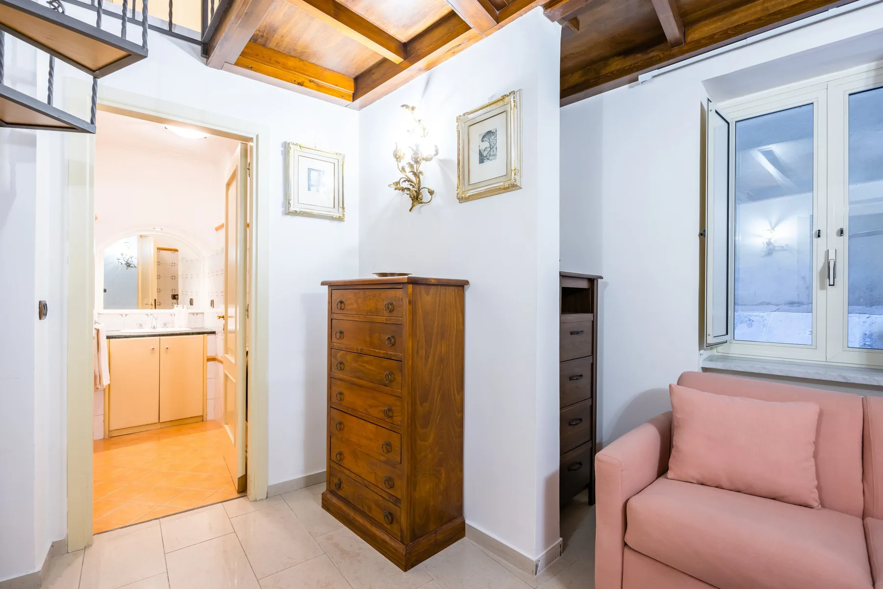 A bright living area with pink seating, a wooden chest, and an open doorway to a bathroom.