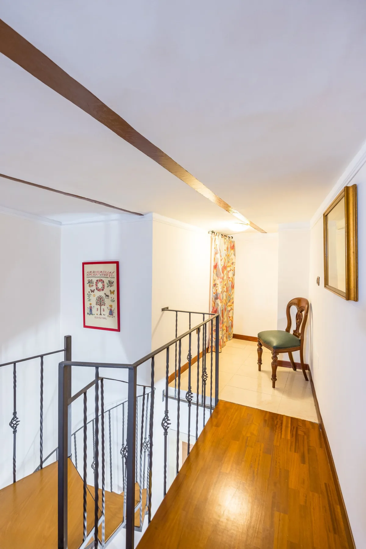 A hallway with a wooden floor, railing, and a chair near a curtained window.