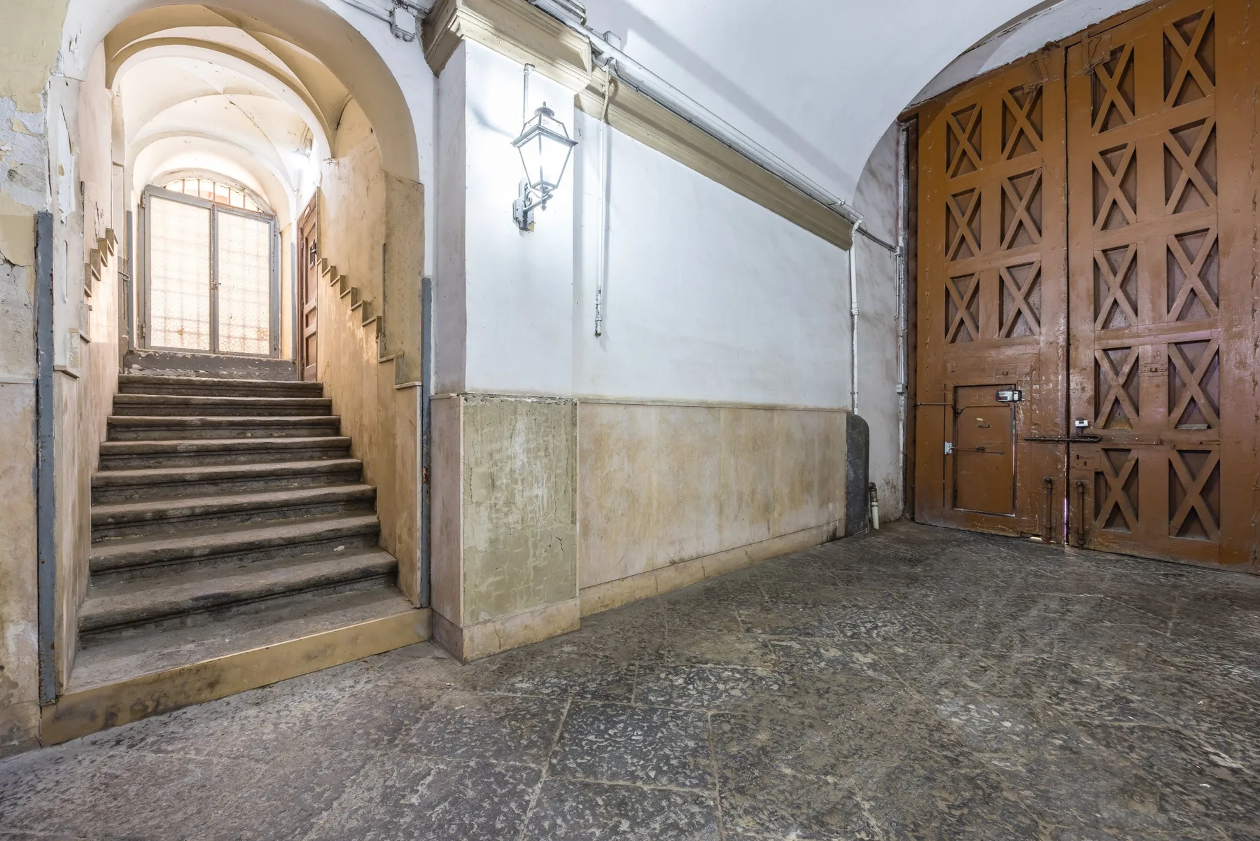 An arched stone corridor with stairs leading to a bright window beside large wooden doors.