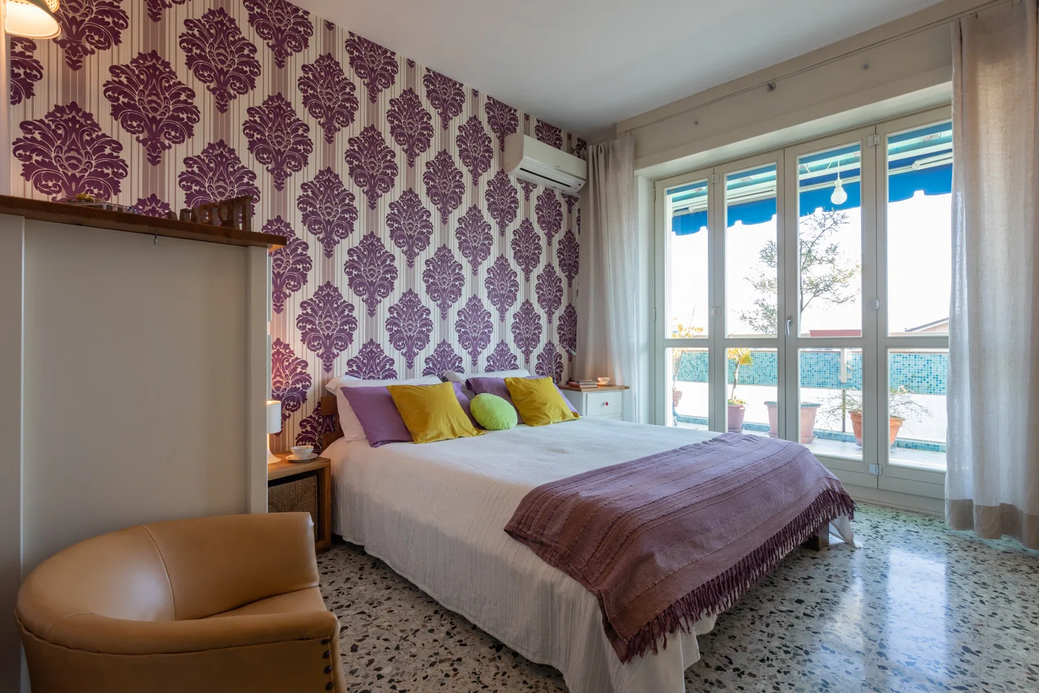 A bedroom with patterned wallpaper and a bed beside large windows.