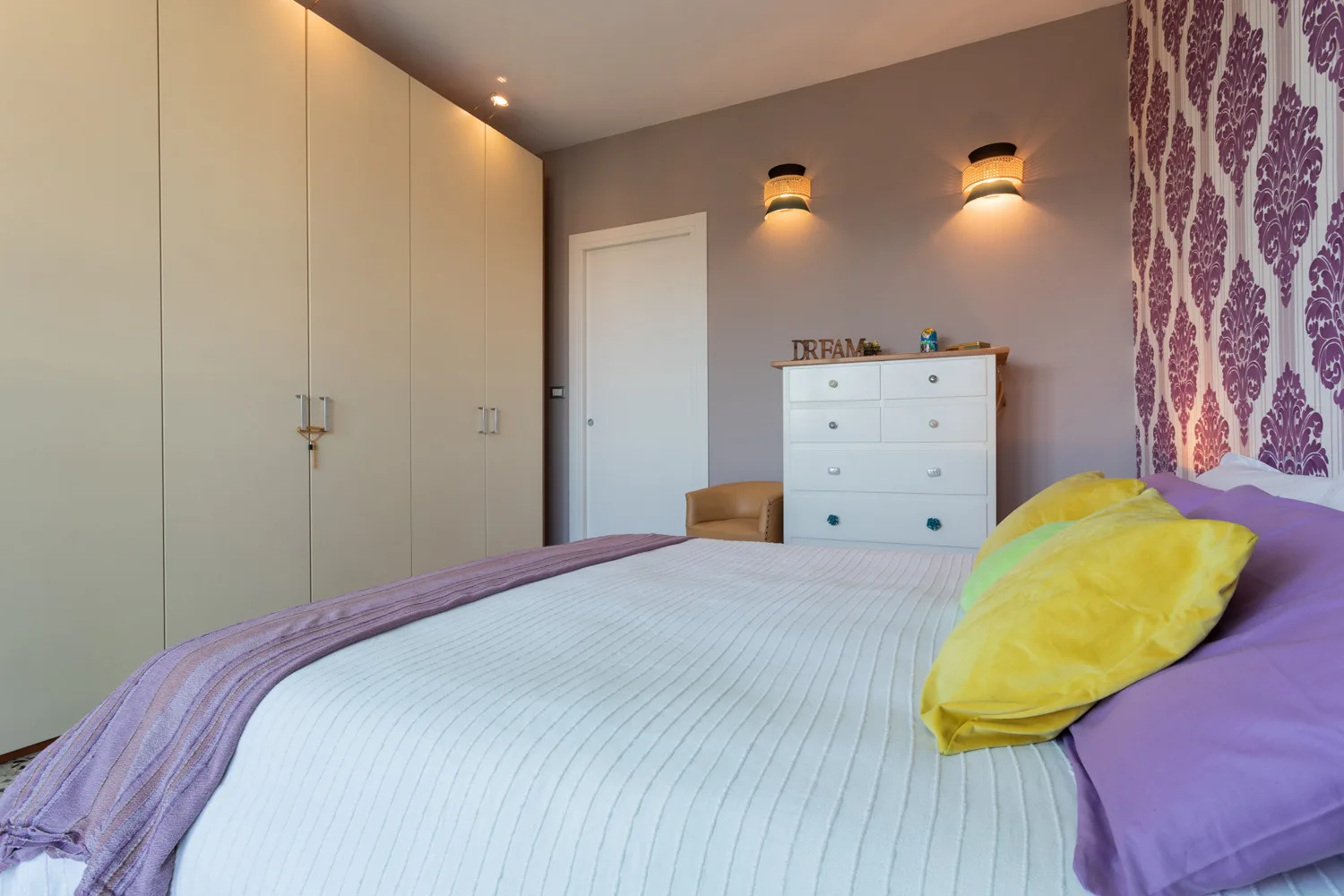 A bedroom with a neatly made bed, white wardrobe, and chest of drawers.