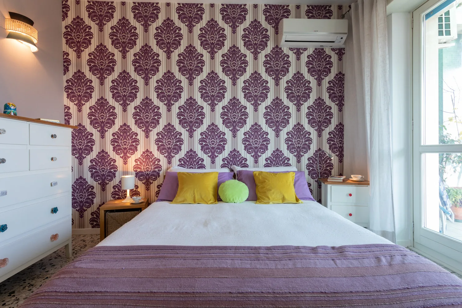 A bedroom with a neatly made bed, patterned wallpaper, and a large window.