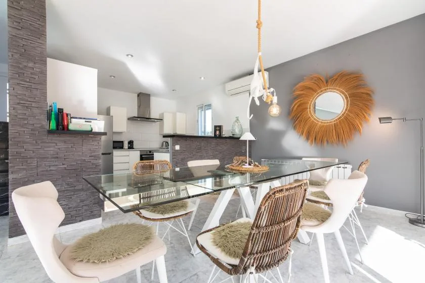 A dining area with a glass table and woven seating beside an open kitchen.