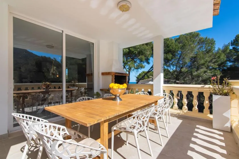 A covered outdoor dining area with a wooden table and white seating overlooks trees.