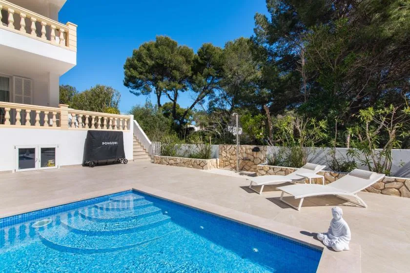 A swimming pool and paved terrace sit beside a white building, with lounge chairs nearby.