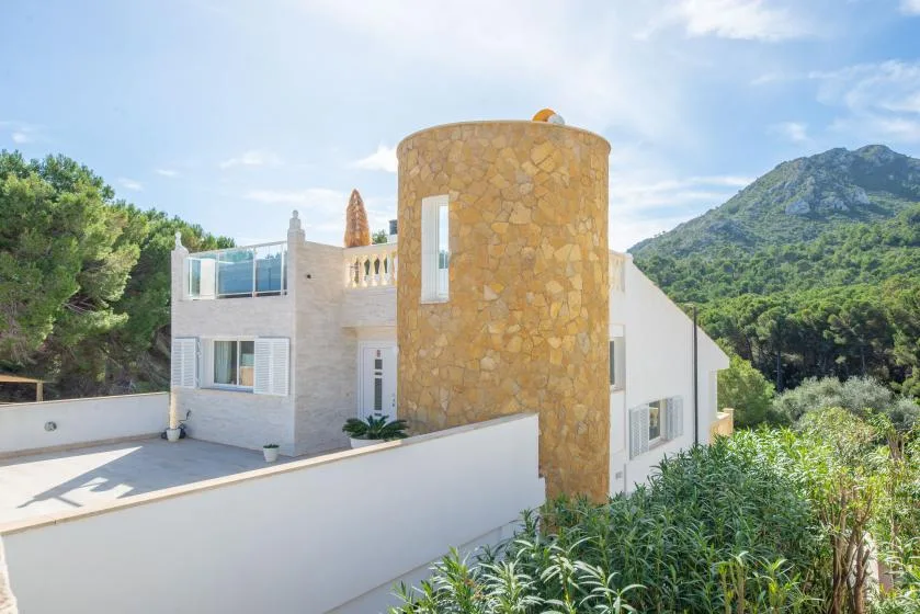 A white house with a rounded stone exterior section stands among greenery beneath a mountain.