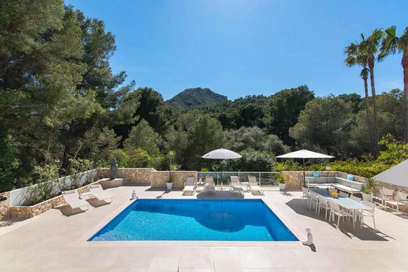 An outdoor swimming pool sits among trees, with lounge seating and umbrellas nearby.