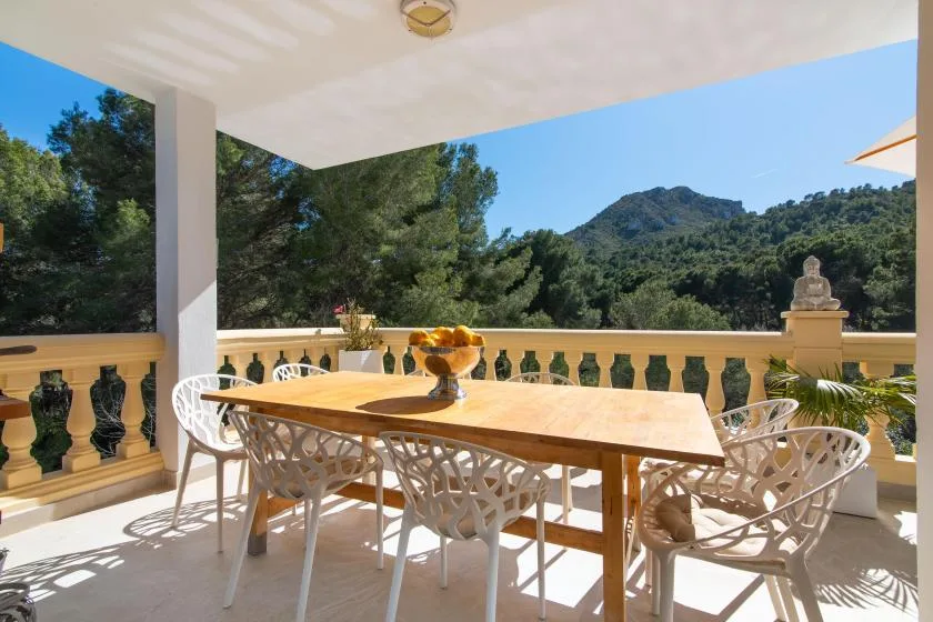A covered terrace with a dining table and seating overlooks wooded hills.