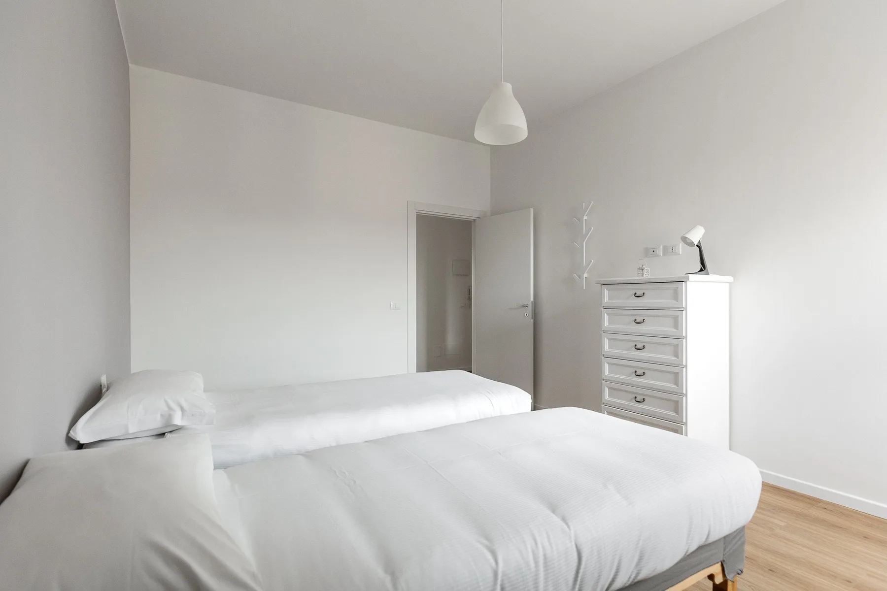 Un dormitorio luminoso con ropa de cama blanca y una cómoda junto a la puerta abierta.
