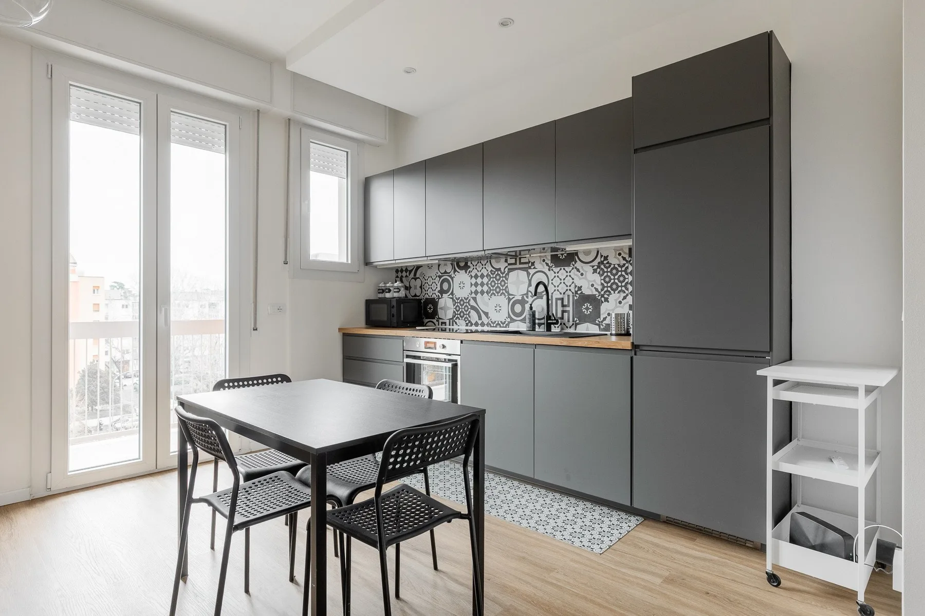 Una luminosa cocina con comedor, gabinetes oscuros y una mesa junto a ventanas altas.