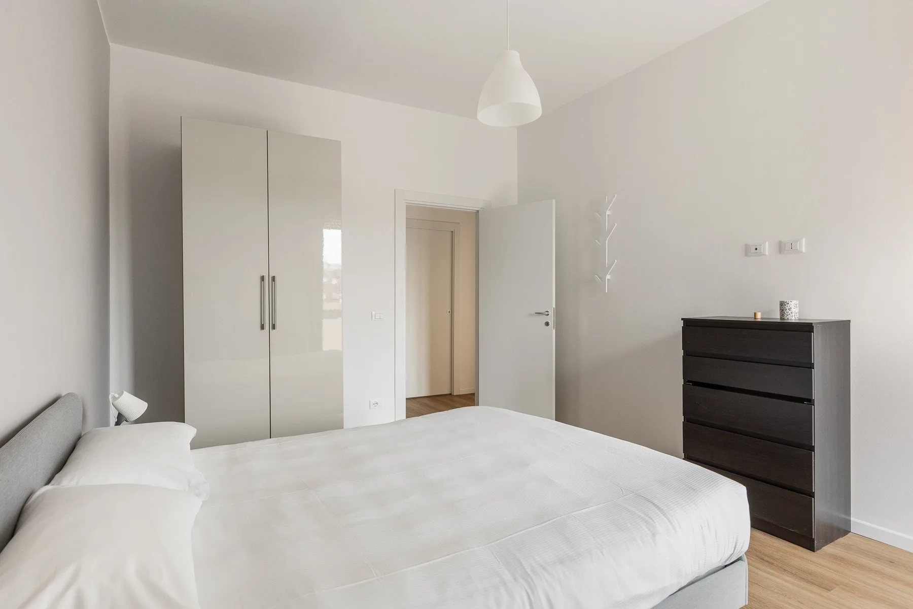 A bright bedroom with a white bed, wardrobe, and dark dresser.