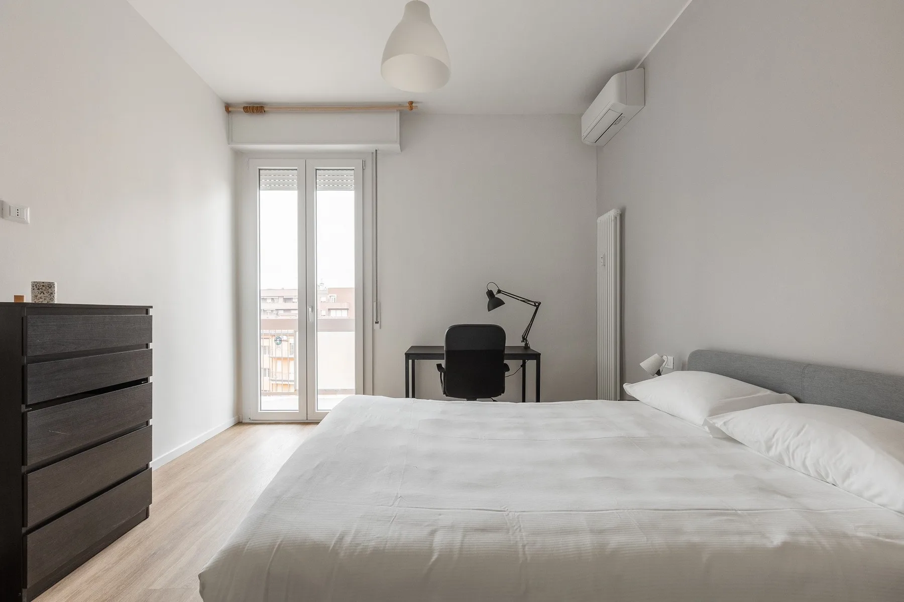 Un dormitorio luminoso con una cama, un escritorio y puertas de cristal que dan a un balcón.