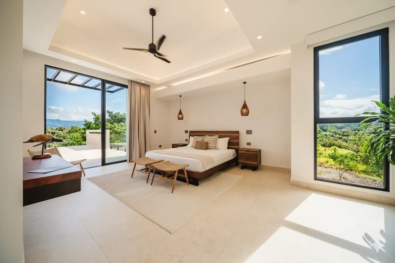 Casa Ama · 15. This bright bedroom features a king-size bed, a tray ceiling, and natural light from large sliding doors leading to a terrace and a window with views of tropical hills.