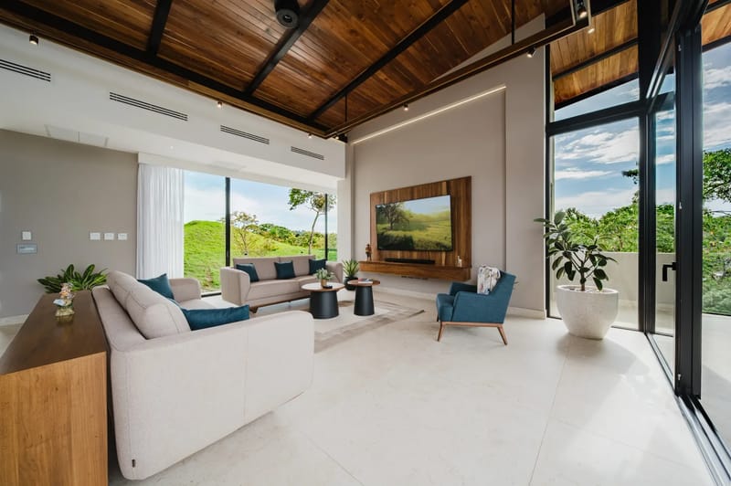 Casa Ama · 27. The living room features a high vaulted wooden ceiling, light tiled floors, and expansive glass doors that merge the interior with views of green hills and trees.