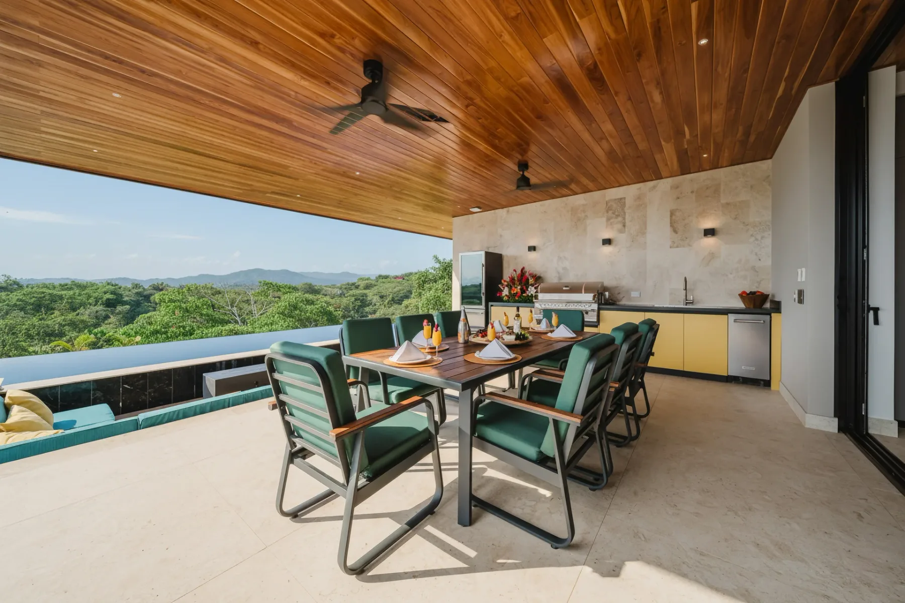 Casa Ama · 28. A spacious covered terrace with a wood-paneled ceiling features an outdoor dining area and kitchen, opening to an infinity pool and expansive views of the tropical landscape.