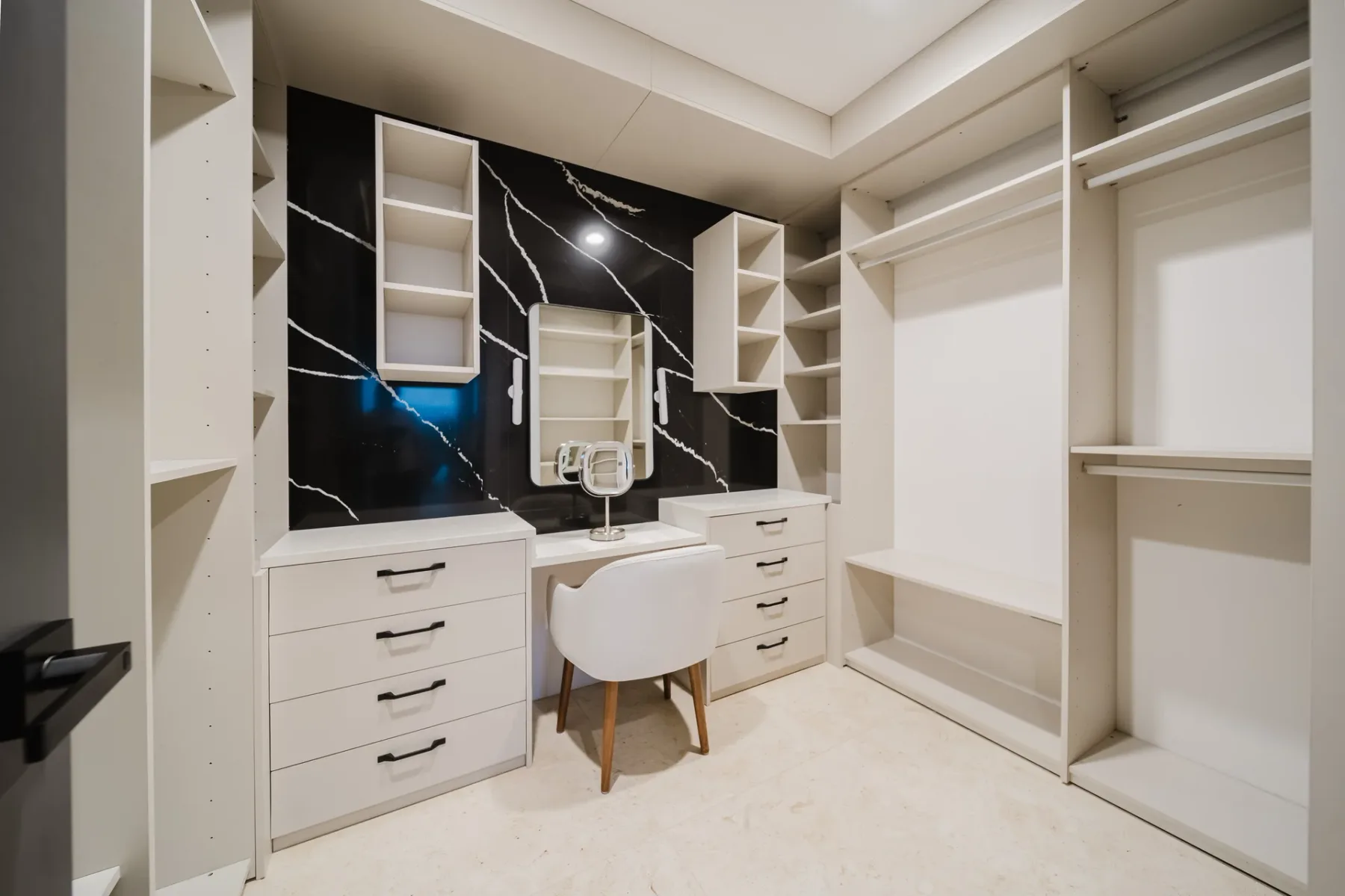 Casa Ama · 38. A dedicated dressing room features extensive built-in light-colored cabinetry and shelving, with a central vanity area set against a striking dark marble-patterned accent wall, illuminated by overhead lighting.