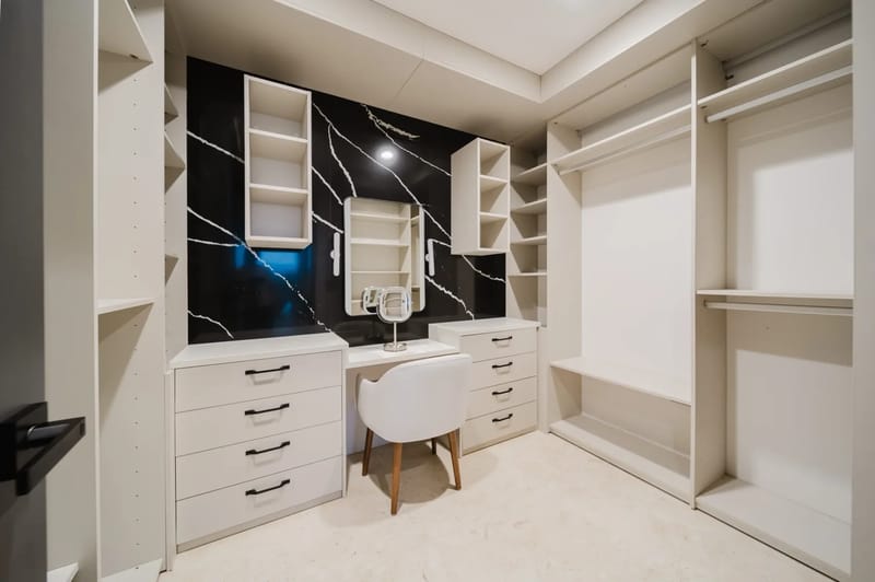 Casa Ama · 38. A dedicated dressing room features extensive built-in light-colored cabinetry and shelving, with a central vanity area set against a striking dark marble-patterned accent wall, illuminated by overhead lighting.