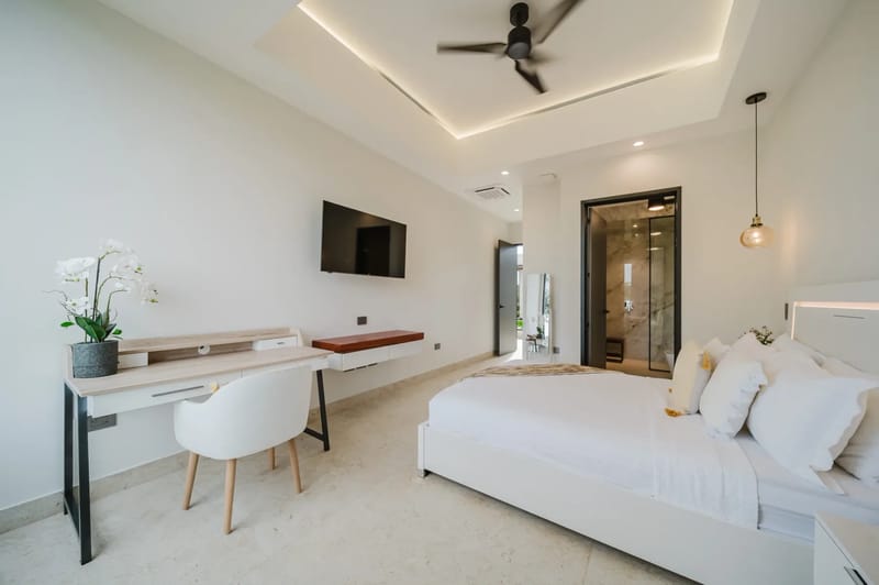 Casa Ama · 8. A bright bedroom features a modern bed with integrated headboard lighting, a desk area, a wall-mounted television, and direct access to an outdoor area and a private bathroom with a visible marble shower.