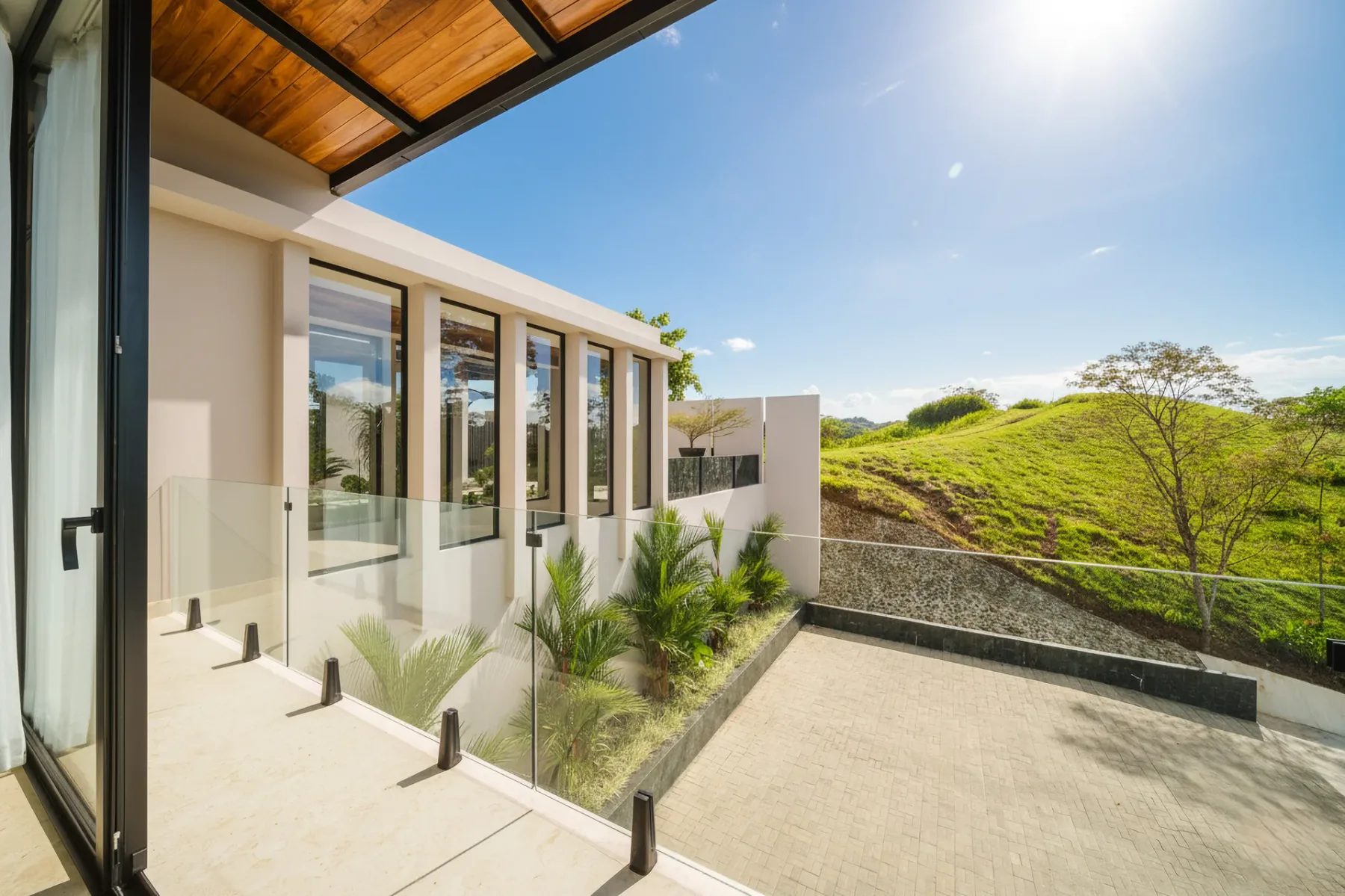 Casa Ama · 26. A covered outdoor terrace features a wooden ceiling and glass railing, offering views of a modern building facade, a paved patio, and sunlit green hills under a clear blue sky.