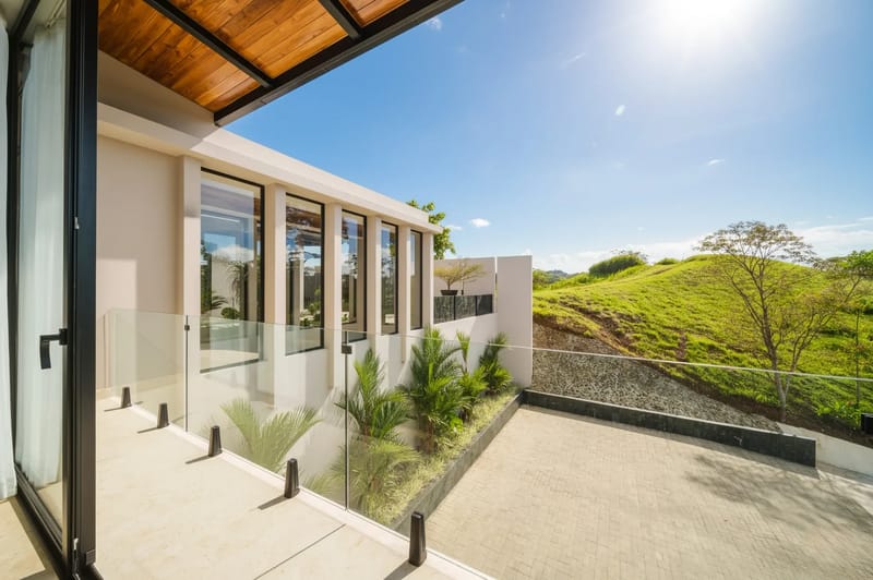 Casa Ama · 26. A covered outdoor terrace features a wooden ceiling and glass railing, offering views of a modern building facade, a paved patio, and sunlit green hills under a clear blue sky.