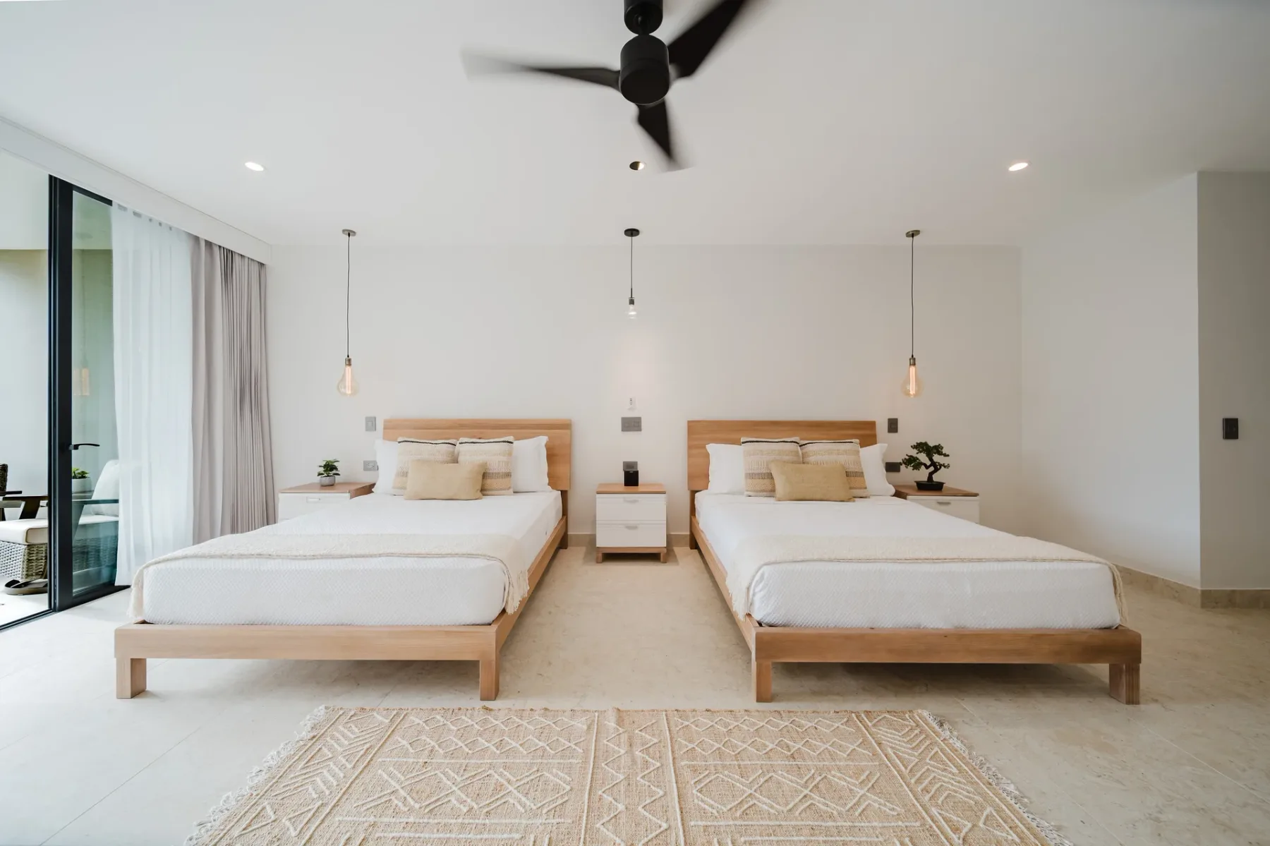 Casa Ama · 25. A bright bedroom features two wooden-framed beds, a light tiled floor, and a large sliding glass door opening to an outdoor area, complemented by a ceiling fan and pendant lighting.