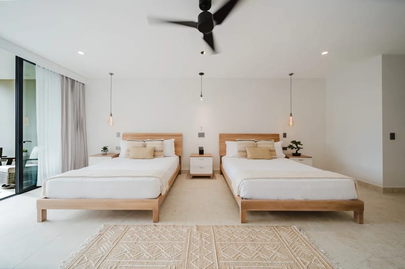Casa Ama · 25. A bright bedroom features two wooden-framed beds, a light tiled floor, and a large sliding glass door opening to an outdoor area, complemented by a ceiling fan and pendant lighting.