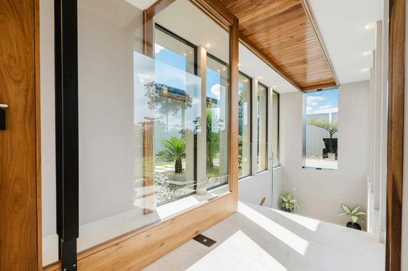 Casa Ama · 20. This bright interior hallway features expansive glass walls with warm wood framing, offering a tranquil garden view and natural light spilling over the light-toned floors towards a descending staircase.