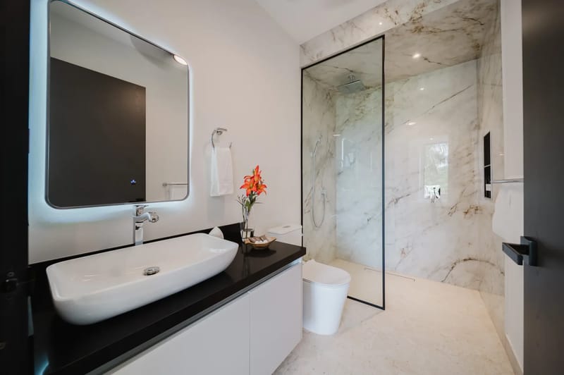 Casa Ama · 45. A contemporary bathroom features a floating vanity with a vessel sink, a backlit mirror, and a walk-in shower with marble-look walls and natural light from an outdoor view.
