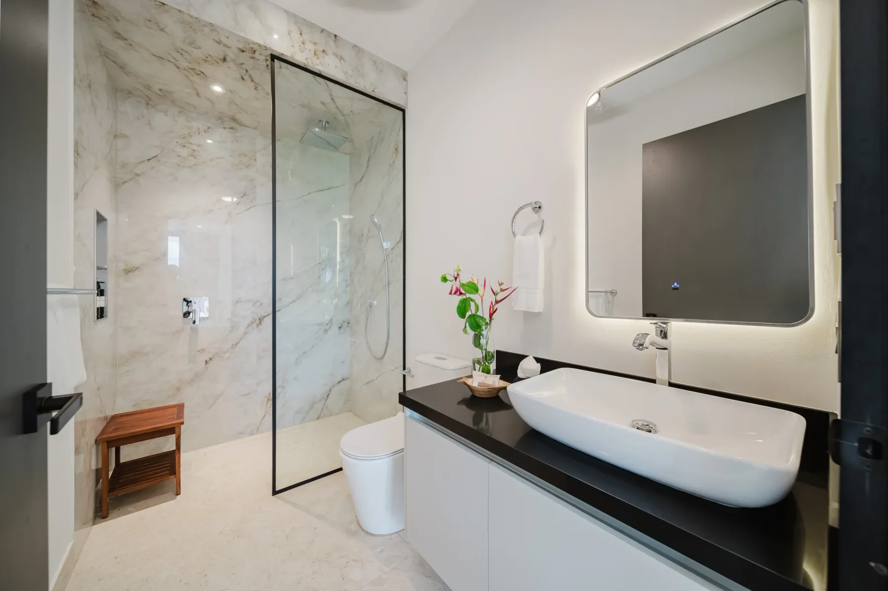 Casa Ama · 24. This modern bathroom features a spacious walk-in shower with marble-look tiling, a glass enclosure, and an integrated wooden bench, alongside a sleek vanity with a vessel sink and a backlit mirror.