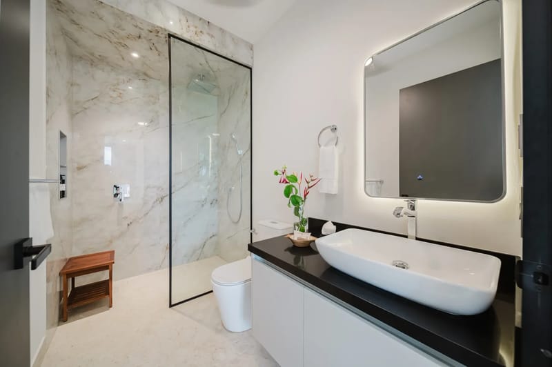Casa Ama · 24. This modern bathroom features a spacious walk-in shower with marble-look tiling, a glass enclosure, and an integrated wooden bench, alongside a sleek vanity with a vessel sink and a backlit mirror.