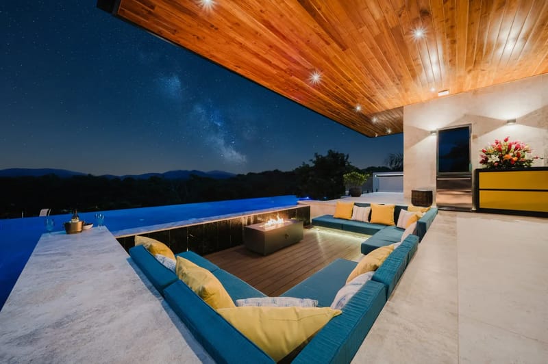 Casa Ama · 50. A covered outdoor terrace with an L-shaped lounge and fire pit overlooks an infinity pool and a night sky filled with stars above distant mountains.