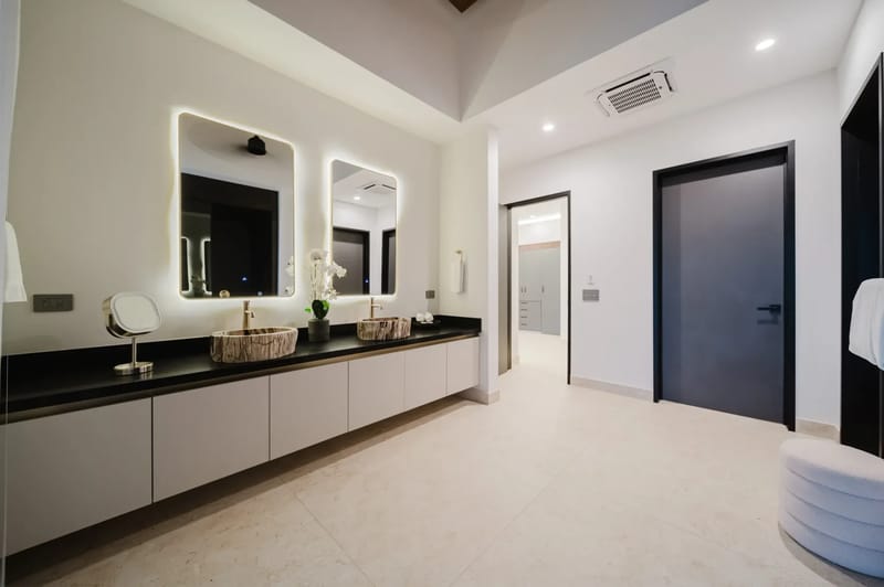 Casa Ama · 21. This spacious bathroom features a double vanity with natural stone vessel sinks, backlit mirrors, ceiling air conditioning, and flush doors leading to adjacent private areas.