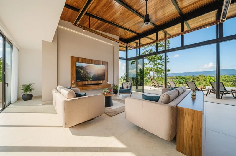 Casa Ama · 44. A bright living room features light-toned sofas, a wooden slatted ceiling with a fan, and floor-to-ceiling glass walls providing panoramic views of green mountains and opening to an outdoor terrace.