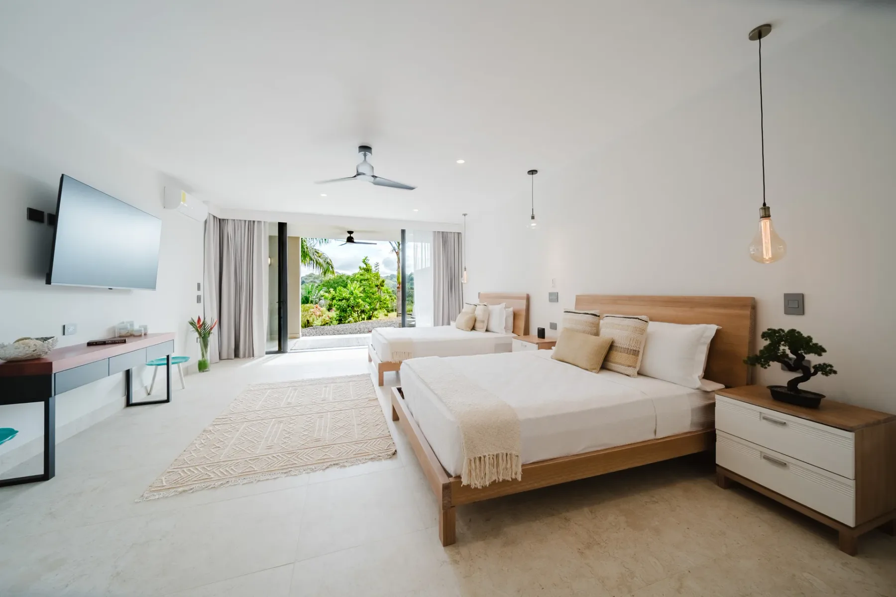 Casa Ama · 32. This airy bedroom, with two beds and light-colored floors, opens fully to a vibrant tropical garden through expansive glass doors, inviting the outdoors in.