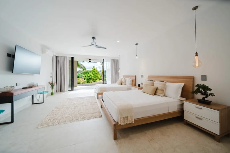Casa Ama · 32. This airy bedroom, with two beds and light-colored floors, opens fully to a vibrant tropical garden through expansive glass doors, inviting the outdoors in.