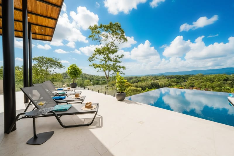 Casa Ama · 33. A tiled terrace with shaded lounge chairs extends to an infinity pool, overlooking a dense green landscape under a bright sky.