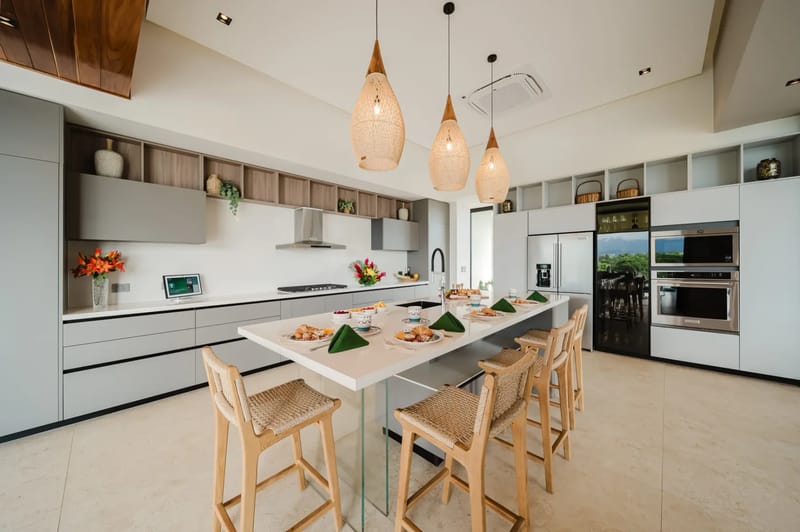 Casa Ama · 31. A bright, contemporary kitchen features light grey cabinetry, a large island with bar seating, integrated appliances, and views to the outdoors.